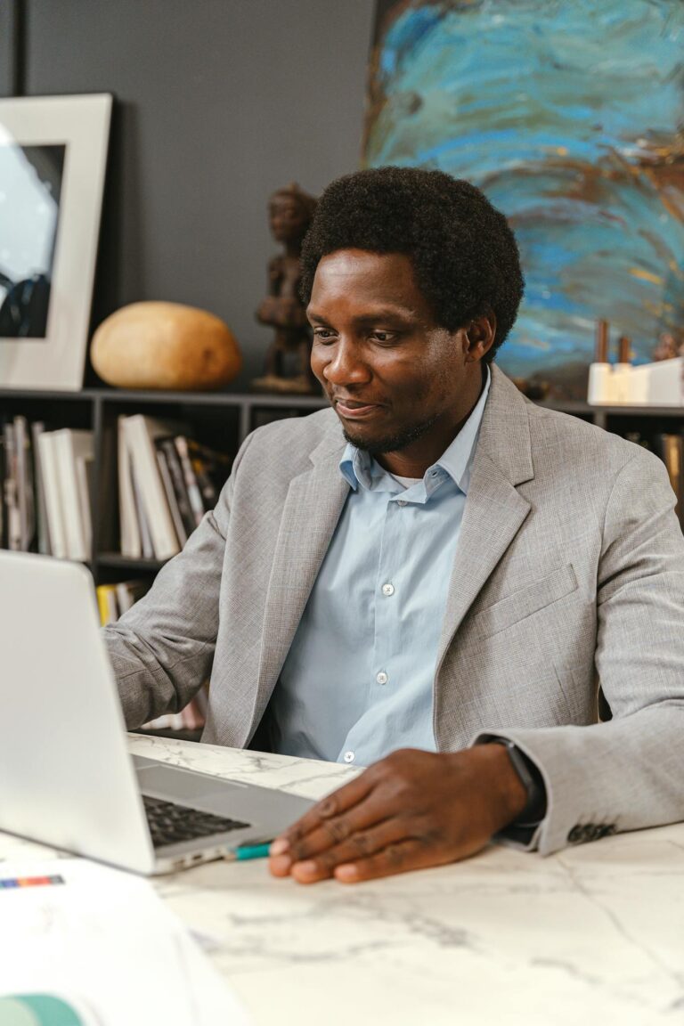 An adult man in a gray suit working on a laptop in an office environment, smiling and engaged.